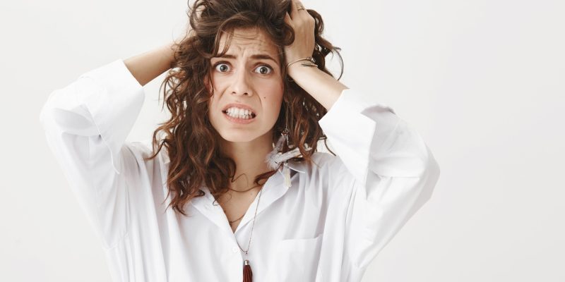 indoor portrait of stunned anxious woman who do not know what to do and having panic attack, holding hands on hair, frowning at camera, standing over gray background. Someone stole her phone.